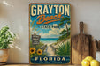 A vintage-style sign for Grayton Beach State Park in Florida, featuring a beach scene with a wooden walkway, palm trees, and a blue sky with clouds.