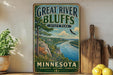 A vintage-style sign for Great River Bluffs State Park in Minnesota, featuring a scenic illustration of the park's landscape with a river, mountains, and wildlife.