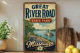 A vintage-style sign for the Great River Road State Park in Mississippi, featuring a scenic illustration of a river, trees, and a steamboat.