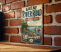 A vintage-style metal sign for the Great River Road State Park in Mississippi, featuring a scenic illustration of a river and a steamboat.