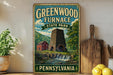 A vintage-style sign for Greenwood Furnace State Park in Pennsylvania, featuring a stone chimney and a red brick building, with a waterfall and lush greenery in the background.