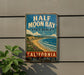 A vintage-style metal sign for Half Moon Bay State Beach in California, featuring a beach scene with mountains and a blue sky.
