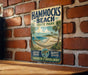 A vintage-style metal sign for Hammocks Beach State Park in North Carolina, featuring a beach scene with palm trees and a hammock.