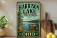 A vintage-style sign for Harrison Lake State Park, featuring a scenic landscape with a lake, trees, and mountains.