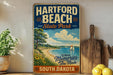 A vintage-style sign for Hartford Beach State Park in South Dakota, featuring a scenic illustration of a beach, sailboat, and mountains.