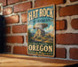 A vintage-style metal sign for the Hat Rock State Heritage Site in Oregon, featuring a large rock formation and the text "Discover Oregon's Past" and "Oregon".