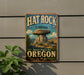 A vintage-style metal sign for the Hat Rock State Heritage Site in Oregon, featuring a large mushroom-shaped rock formation against a scenic background.