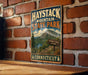 A vintage metal sign for Haystack Mountain State Park in Connecticut, featuring a scenic mountain landscape and trail sign.