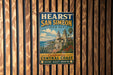 A vintage metal sign for Hearst San Simeon State Park, featuring a scenic illustration of a coastal town with palm trees and mountains.