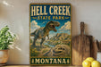 A vintage-style metal sign for Hell Creek State Park in Montana, featuring a dinosaur and a scenic landscape.