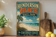 A vintage-style sign for Henderson Beach State Park in Florida, featuring a beach scene with palm trees and a clear blue sky.