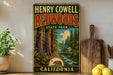 A vintage-style metal sign for Henry Cowell Redwoods State Park in California, featuring a scenic illustration of the park's landscape with towering redwood trees, a stream, and a bridge.