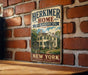 A vintage-style metal sign for Herkimer Home, a historic site in New York, is displayed on a brick wall.