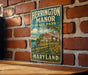 A vintage-style metal sign for Herrington Manor State Park in Maryland, featuring a house and a tree, is displayed on a brick wall.