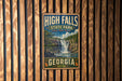 A vintage metal sign for High Falls State Park in Georgia, featuring a waterfall and the text "High Falls State Park, Explore More Discoveries, Georgia".
