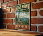 A vintage-style metal sign for High Point State Park in New Jersey, featuring a scenic landscape with a monument and a river.