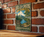 A vintage-style metal sign for Hills Creek State Park in Pennsylvania, featuring a scenic landscape with a river, mountains, and a picnic area.