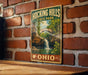 A vintage-style metal sign for Hocking Hills State Park in Ohio, featuring a scenic image of a waterfall and lush greenery.