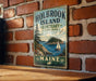 A vintage-style metal sign for Holbrook Island, Maine, featuring a scenic illustration of a sailboat on the water and a forested island in the background.