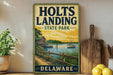 A vintage-style metal sign for Holts Landing State Park in Delaware, featuring a scenic illustration of a lake, dock, and house.