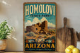 A vintage-style sign for Homolovi State Park in Arizona, featuring an illustration of a desert landscape with a large stone structure and a decorative pot.