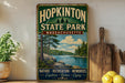 A vintage-style metal sign for Hopkinton State Park in Massachusetts, featuring a scenic landscape with pine trees, a lake, and mountains.