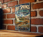 A vintage-style metal sign for Hot Springs State Park in Wyoming, featuring a scenic image of the natural hot springs and mountains.
