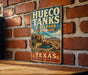 A vintage-style metal sign for Hueco Tanks State Park & Historic Site in Texas, featuring a desert landscape with a small lake and mountains in the background.