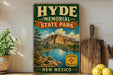 A vintage-style sign for Hyde Memorial State Park in New Mexico, featuring a scenic landscape with mountains, a lake, and a wooden paddleboard.