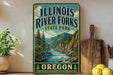 A vintage-style sign for Illinois River Forks State Park, featuring a scenic illustration of a river surrounded by mountains and pine trees.