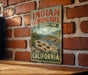 A vintage-style metal sign for Indian Grinding Rock State Historic Park in California, featuring a scenic landscape with mountains and a river.