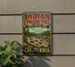 A vintage-style sign for Indian Grinding Rock State Historic Park in California, featuring a scenic landscape with mountains, a river, and a large hole in the ground.