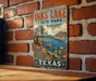 A vintage-style metal sign for Inks Lake State Park in Texas, featuring a scenic landscape with a lake, mountains, and cacti.