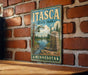 A vintage-style metal sign for Itasca State Park in Minnesota, featuring a scenic landscape with a river, pine trees, and a monument.