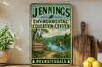 A vintage-style sign for the Jennings Environmental Education Center in Pennsylvania, featuring a scenic landscape with a river, trees, and a bird.