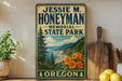 A vintage-style sign for Jessie M. Honeyman Memorial State Park in Oregon, featuring a scenic landscape with pine trees, mountains, and a river.