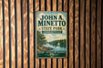 A vintage metal sign for John A. Minetto State Park in Connecticut, featuring a scenic landscape with a lake, trees, and a sailboat.