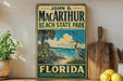 A vintage-style sign for John D. Mac Arthur Beach State Park in Florida, featuring a beach scene with palm trees, a heron, and a blue sky with clouds.