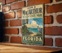 A vintage-style metal sign for John D. Mac Arthur Beach State Park in Florida, featuring a beach scene with palm trees and a bird, is displayed on a brick wall.