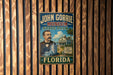 A vintage-style metal sign for the John Gorrie Museum in Florida, featuring an illustration of a man in a suit and tie standing in front of a building with a palm tree.