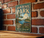 A vintage-style metal sign for John Hens Park in Wisconsin, featuring a scenic landscape with a lake and trees.