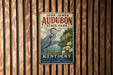 A vintage-style metal sign for John James Audubon State Park in Kentucky, featuring a blue heron and a scenic landscape.