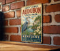 A vintage-style metal sign for John James Audubon State Park in Kentucky, featuring a large blue heron and a scenic landscape.