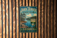 A vintage-style metal sign for John Tanner State Park in Georgia, featuring a scenic landscape with a lake and pine trees.