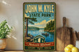 A vintage-style sign for John W. Kyle State Park, featuring a scenic image of a lake surrounded by pine trees and mountains.