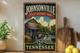 A vintage-style sign for Johnsonville State Historic Park in Tennessee, featuring a wooden building and a waterfall, is displayed on a shelf next to a wooden cutting board and some lemons.
