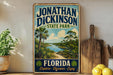 A vintage-style sign for Jonathan Dickinson State Park in Florida, featuring a scenic landscape with palm trees and a river.