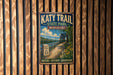 A vintage-style metal sign for the Katy Trail in Missouri, featuring a scenic illustration of a bridge over a river and a path leading through a wooded area.