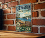 A vintage metal sign for Kewaydin State Park, featuring a scenic illustration of a lake, mountains, and a sailboat, mounted on a brick wall.