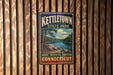 A vintage metal sign for kettletown state park hangs on a wooden wall, featuring a scenic image of a lake surrounded by trees and mountains.